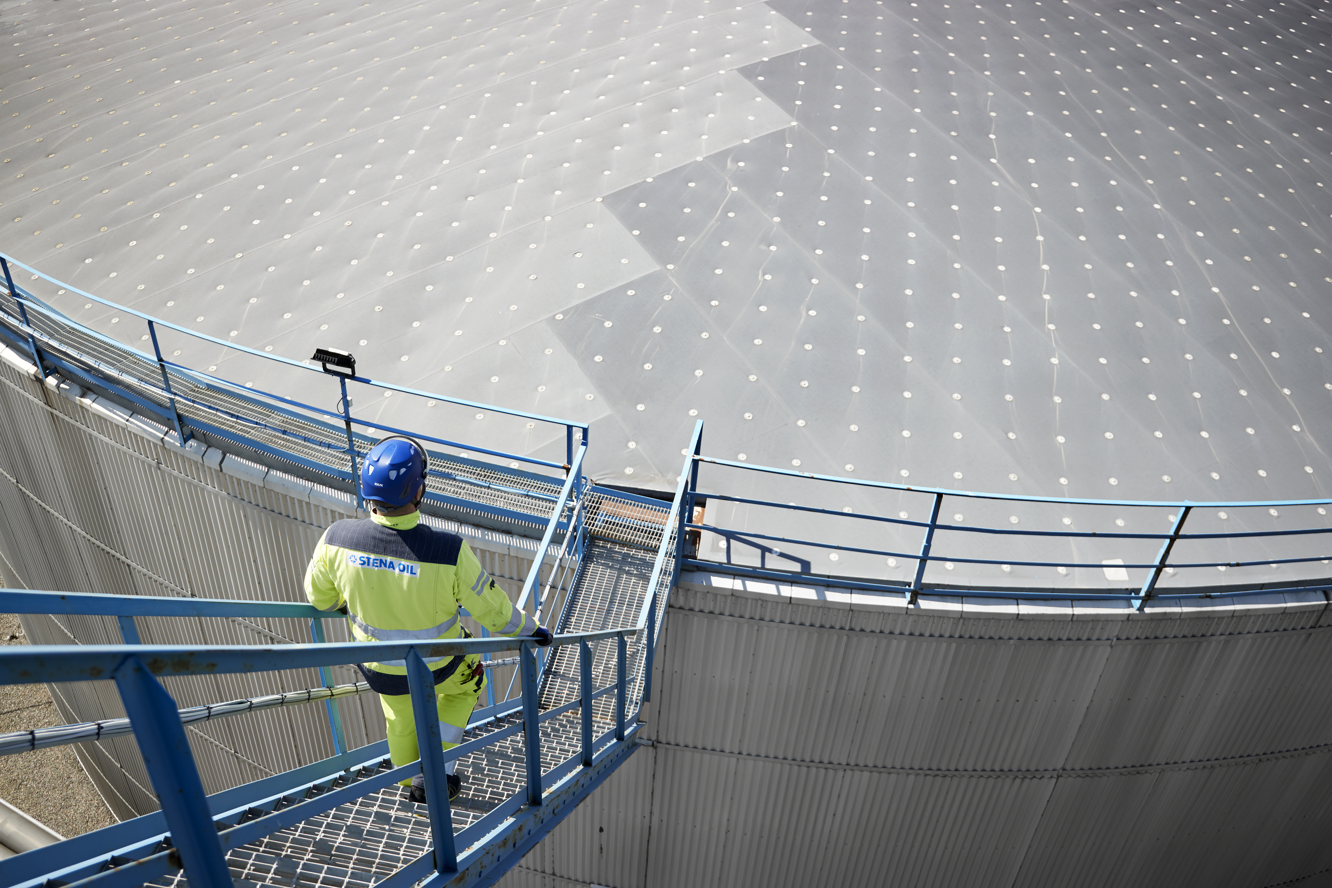 Man in yellow jacket and construction helmet walking down stairs to an oil plant